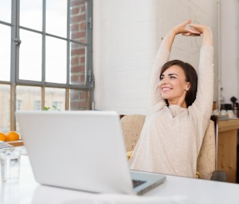 Happy-relaxed-young-woman-sitting-in-her-kitchen-with-a-laptop-in-front-of-her-stretching-her-arms-above-her-head-and-looking-out-of-the-window-with-a-smile